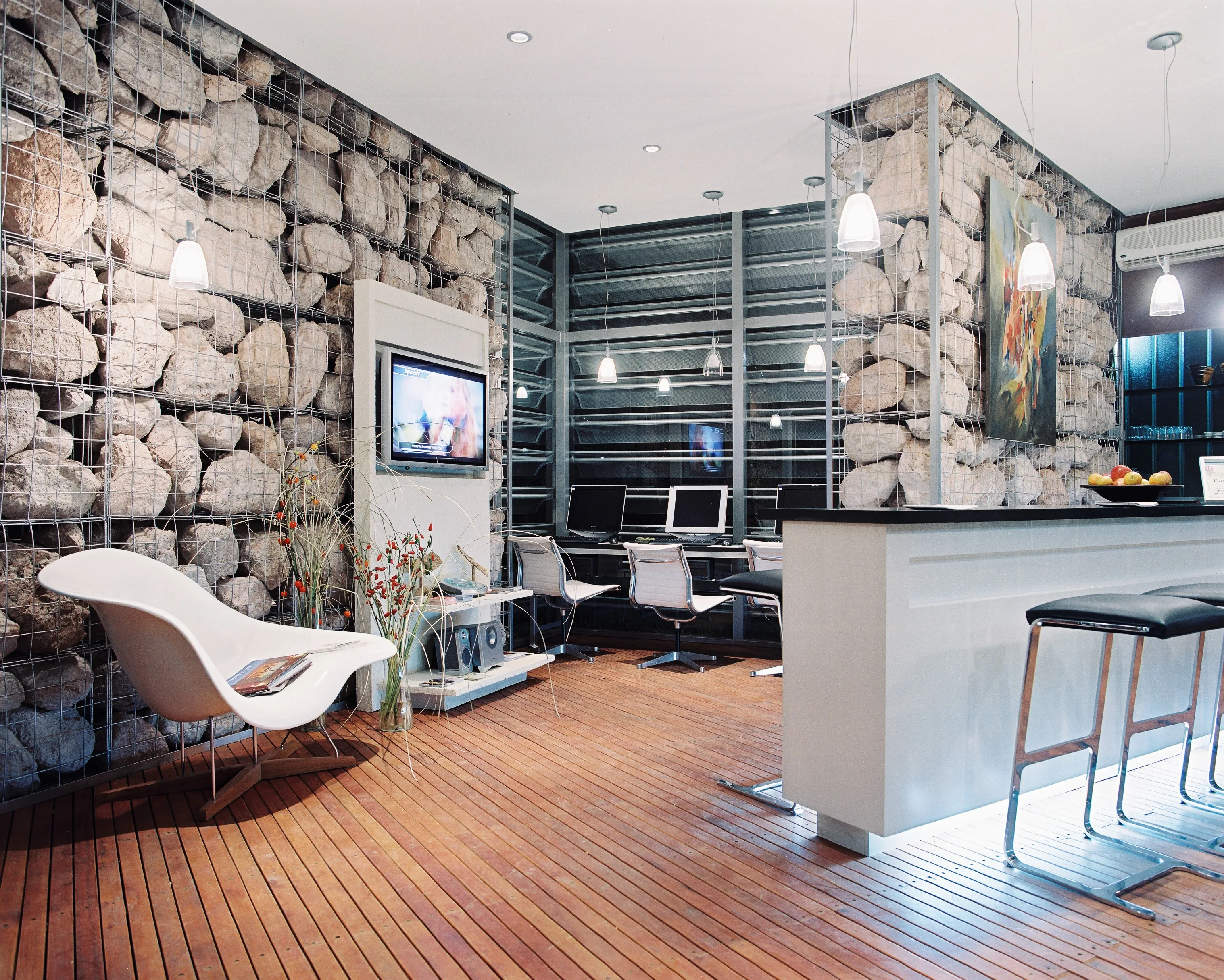 Interior del estudio Parrado Arquitectura en Pilará, con pared de piedra tipo gabión, piso de madera y sillas Eames